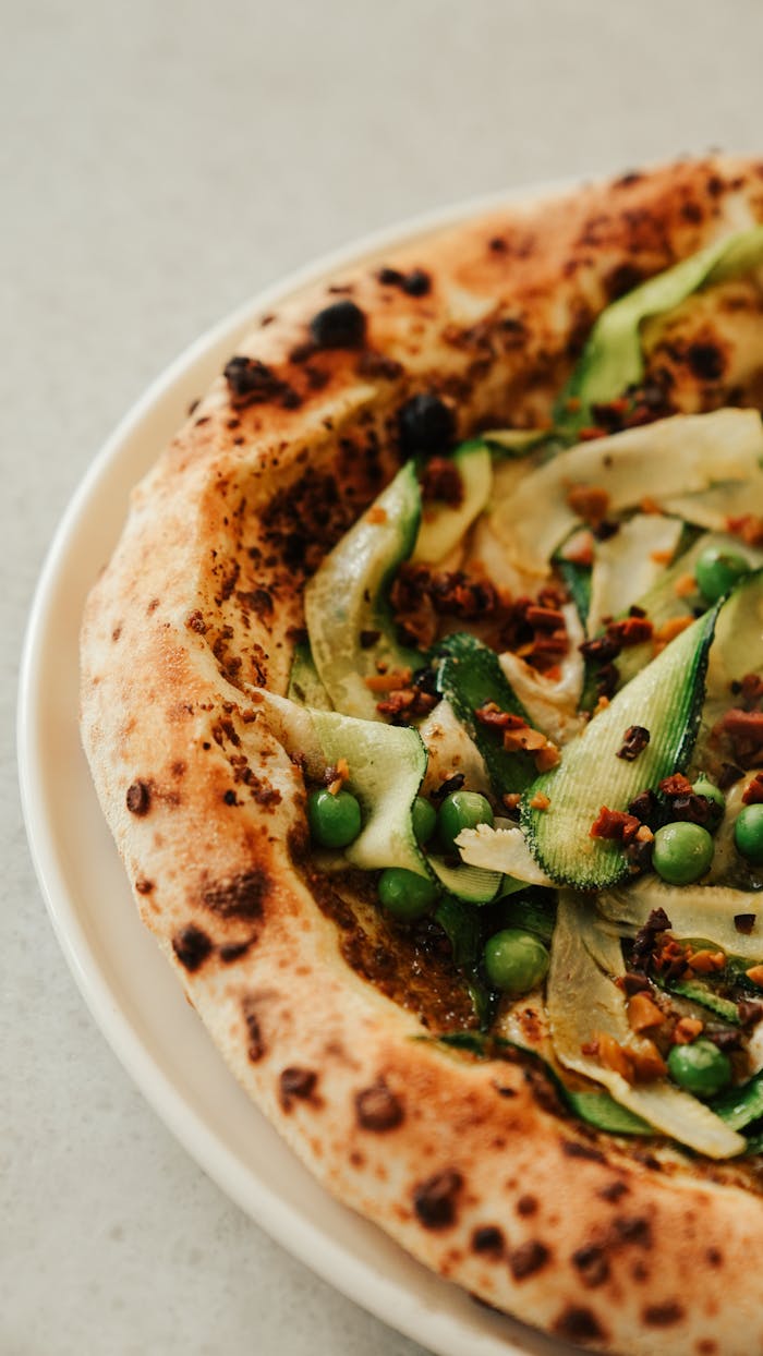 Close-up of a delicious gourmet pizza topped with fresh green peas, zucchini, and crispy bits on a golden crust.