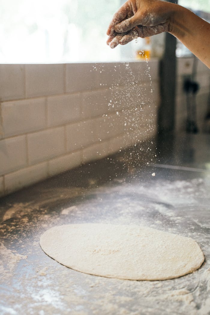 services-img A close-up of a hand sprinkling flour over dough on a kitchen counter in an indoor setting.