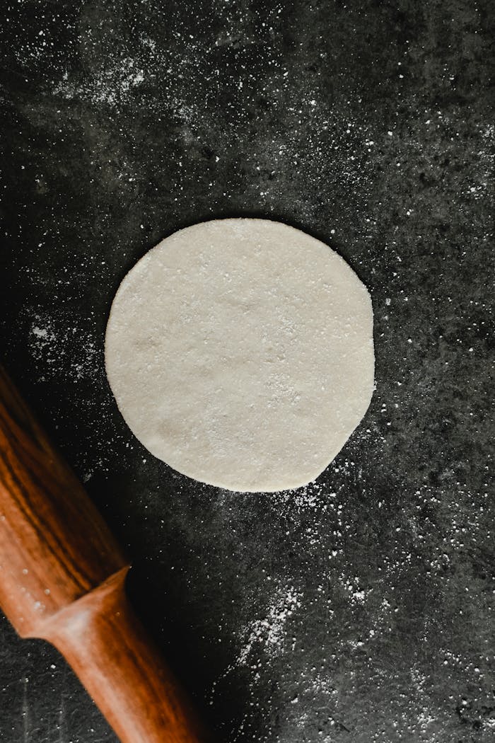 services-01 Close-up of round dough on dark surface with rolling pin, showing texture.